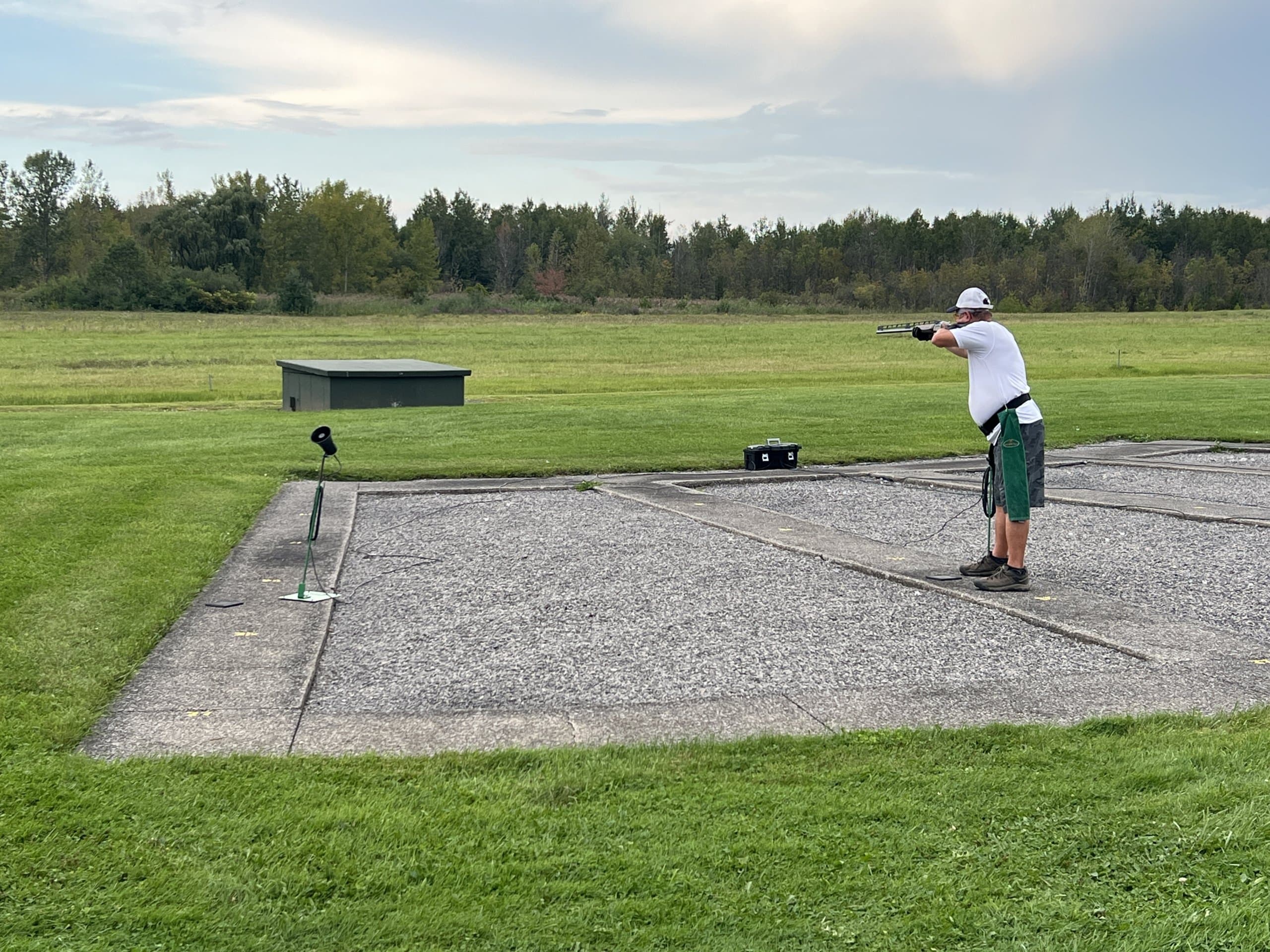 A trapshooter on the firing line behind the trap house on a green New England field